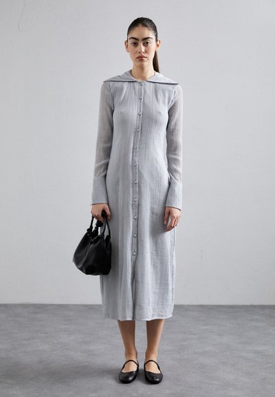Light gray button-up dress with long sleeves, textured fabric, hooded collar, and side slits; paired with black handbag and flats.