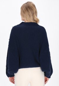 Pulover tricotat de culoare bleumarin, cu guler înalt, mâneci tip balon și textură ribbată. Prezintă nasturi aurii pe umăr.