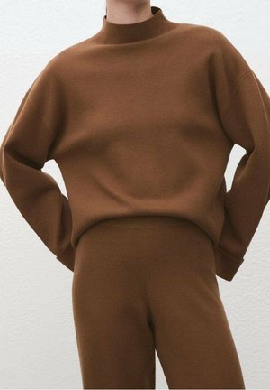 Personne portant un haut à manches longues marron et un pantalon assorti marron, sur un fond clair uni, les mains posées sur les hanches.