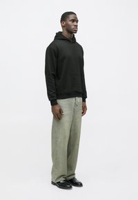 Μαύρη φούτερ μπλούζα με κουκούλα και μπροστινή τσέπη, σε συνδυασμό με ανοιχτό πράσινα φαρδιά τζιν και μαύρα παπούτσια. Απλή, χαλαρή γραμμή, λεία υφή.