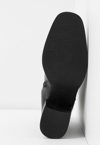 Sól černého kotníkového boty s texturovaným podpatkem, stojící kolmo na bílé rohové zdi a podlaze.