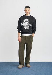 Μαύρο φούτερ με λευκό γραφιστικό σχέδιο κοχυλιού και βίδας, συνδυασμένο με ελιάς πράσινο παντελόνι με φαρδιά πόδια και παπούτσια ταουπέ.