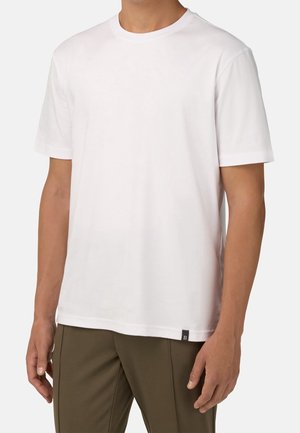 Hombre con camiseta sencilla blanca de manga corta y cuello redondo, combinada con pantalones verde oliva, posando frente a un fondo liso.