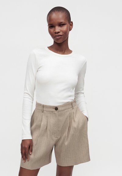 Femme portant un haut blanc à manches longues et un short beige à fines rayures taille haute avec plis et fermeture par bouton.