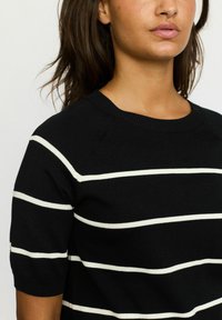 Black knit sweater with white horizontal stripes, short sleeves, and a rounded neckline. Features textured ribbing at the neck.