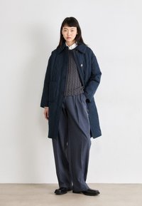 Manteau long matelassé bleu marine avec un col pointu, associé à un pull en maille torsadée gris et un pantalon gris ample ; chaussures noires élégantes.