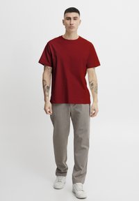 Red cotton t-shirt with short sleeves, paired with light gray tailored pants and white sneakers. Features visible tattoos on arms.