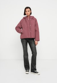 Μονωμένο μπουφάν σε μπορντό χρώμα με κουκούλα, κουμπιά με κλικ και άνετη εφαρμογή, συνδυασμένο με σκούρα τζιν και μαύρα αθλητικά παπούτσια. Ομαλή υφή και ελάχιστες λεπτομέρειες.