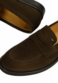 Paire de mocassins en daim marron foncé avec semelles noires et petit logo métallique sur la bride latérale, montrant la doublure intérieure couleur cuir.