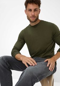 Dunkelgrünes Langarmshirt mit Rundhalsausschnitt, kombiniert mit grauen Jeans. Das Shirt hat eine glatte Textur und eine taillierte Passform.