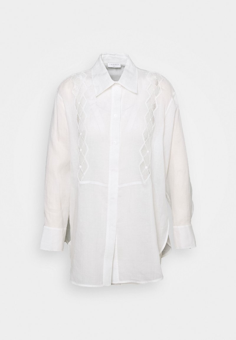 Blouse blanche à manches longues avec col pointu et détails en dentelle transparente en zigzag sur les panneaux avant.