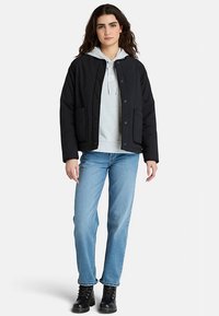 Černá prošívaná bunda se zapínáním na patentky a bočními kapsami, nošená přes šedou mikinu s kapucí, v kombinaci s modrými džínami a černými šněrovacími botami.