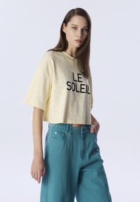 Crop-Top in Hellgelb mit schwarzem Text "LE SOLEIL" und kleinem gepunktetem Muster; kombiniert mit taillenhohen türkisfarbenen, weit geschnittenen Hosen.