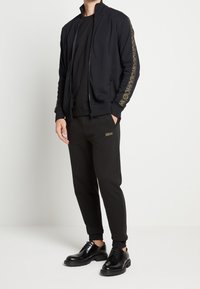 Chaqueta deportiva negra con cremallera, puños acanalados; camiseta negra debajo; pantalones jogger negros con logo; zapatos negros con cordones.