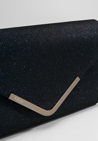 Petit sac pochette rectangulaire noir pailleté avec un accent métallique argenté sur le rabat, détail d'angle en biais, nom de la marque sur la pièce métallique.