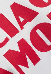 Weiße Stoff mit großen, auffälligen roten Buchstaben und einem strukturierten Druck. Die Buchstaben haben scharfe Kanten und unregelmäßige Abstände, was ein dynamisches Erscheinungsbild erzeugt.