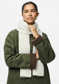 Écharpe blanche à côtes portée sur une veste olive avec des accents de poignet marron. L'écharpe a une texture douce et épaisse, offrant de la chaleur.