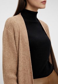 Beige geripptes Cardigan über einem schwarzen Rollkragenpullover, mit langen Ärmeln und offenem Vorderteil, aus einem weichen Material und in legerem Schnitt.