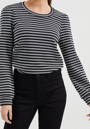 Kvinde iført sort og hvid langærmet trøje med horisontale striber og sorte bukser foran ensartet baggrund.