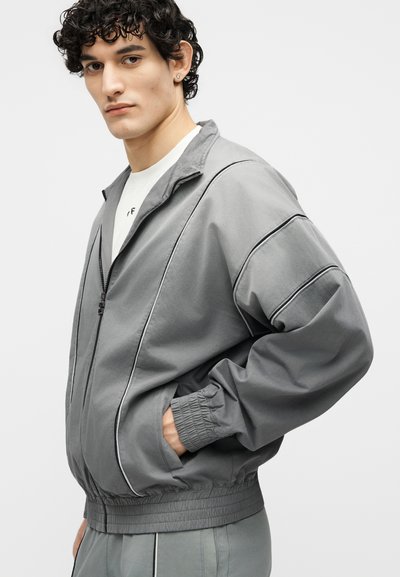 Νέος άνδρας με κατσαρά μαλλιά που φοράει γκρι μπουφάν με φερμουάρ, μαύρες ραφές και ελαστικές μανσέτες, με τα χέρια στις τσέπες, κοιτάζοντας προς τα αριστερά.