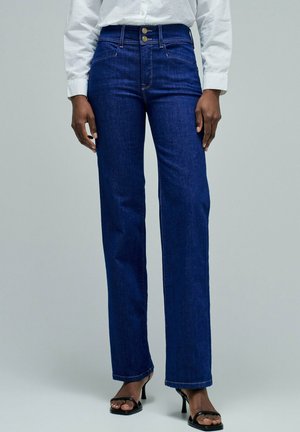 Persona vistiendo jeans de talle alto y pierna recta en azul oscuro, camisa blanca de manga larga y sandalias negras de tacón con punta abierta.