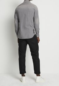 Camicia grigia a maniche lunghe con bottoni, effetto sbiadito, con i polsini arrotolati e un dettaglio a freccia nera sul retro, abbinata a pantaloni neri e scarpe bianche.