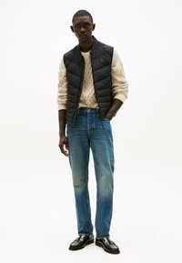 Chaleco acolchado negro sobre un suéter de punto de cable color crema, combinado con jeans azul claro y mocasines negros. Un estilo de capas sencillo y casual.