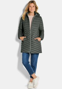 Groene gewatteerde lange jas met capuchon, voorzien van een rits aan de voorkant en zijzakken, gecombineerd met blauwe jeans en witte sneakers.