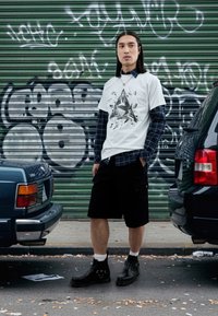 Pantaloni scurți negri, tricou alb cu grafică purtat peste o cămașă în carouri închise la culoare. Ghete negre până la gleznă cu catarame. Cadru urban cu fundal de graffiti.