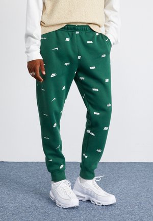 Person iført grønne Nike joggingbukser med hvide logoer og hvide sneakers, der står på et blåt tæppe.