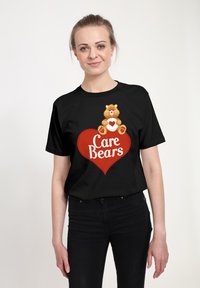 Černé tričko s kresleným medvědem se srdcem, umístěným nad nápisem "Care Bears" bílými písmeny, na pozadí červeného srdce.