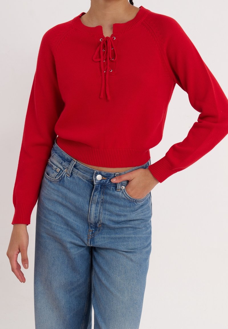 Femme portant un pull en maille rouge à manches longues avec un détail de laçage à l'avant et un jean bleu clair taille haute, main dans la poche.