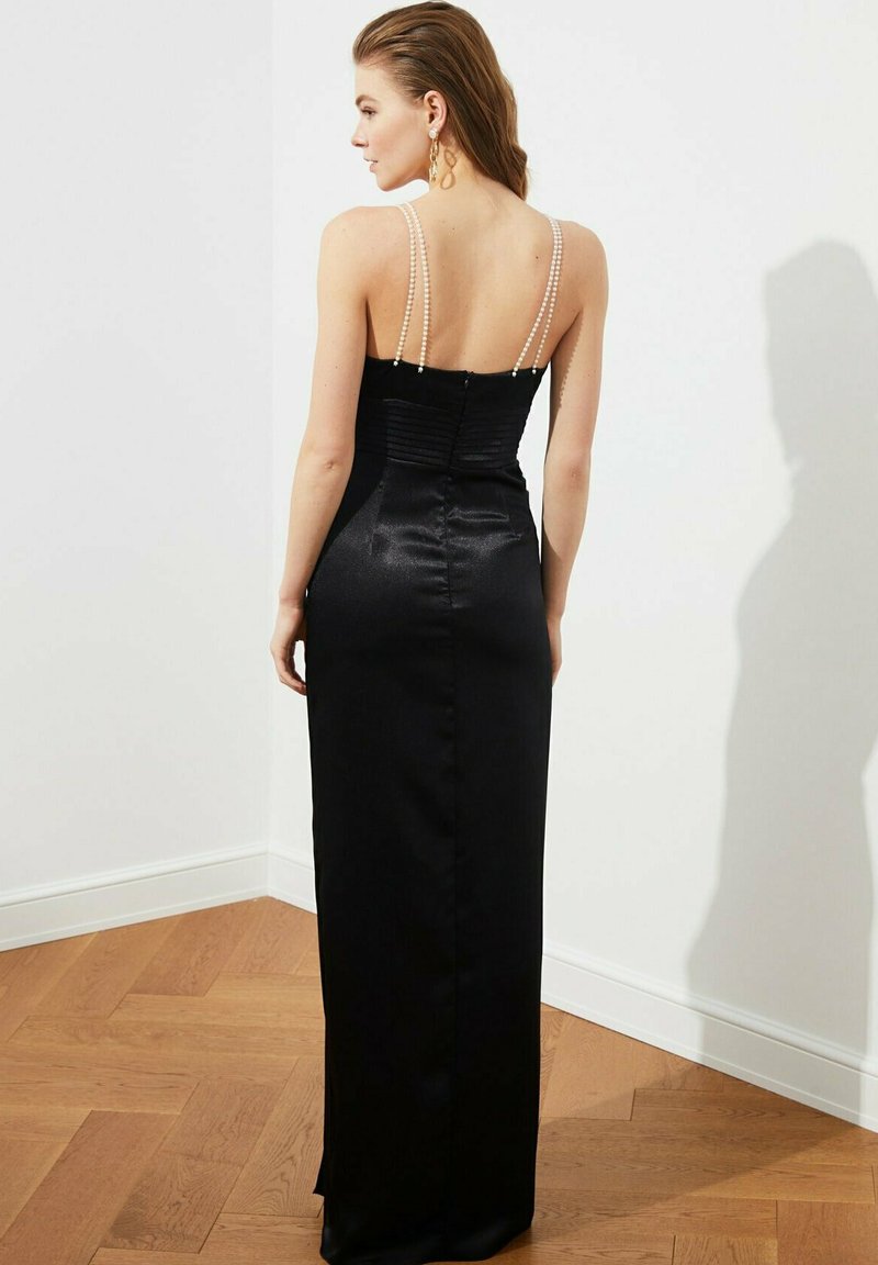Czarna, satynowa suknia wieczorowa o dopasowanej sylwetce, z ramiączkami ozdobionymi perłami i marszczonym tyłem, sięgająca do połowy łydki noszącej ją osoby.