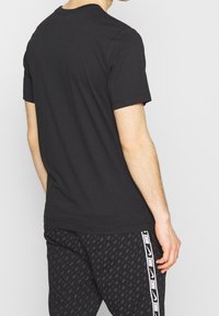 Černé bavlněné tričko s kulatým výstřihem a krátkými rukávy, s jednoduchým designem. Nosené s vzorovanými černými kalhotami s bílým logem.