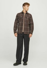 Bruine geruite shirtjack met twee borstzakken, zwarte knopen en een relaxte pasvorm, gelaagd over een beige shirt en gecombineerd met zwarte broeken.