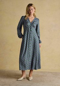 Šaty s dlhými rukávmi v purpurovom a zelenom geometrickom vzore, s hlbokým výstrihom do V, zapínaním na gombíky a voľným strihom. V kombinácii s krémovými šľapkami.