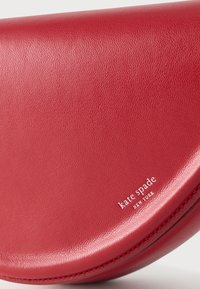 Κόκκινο δερμάτινο τσαντάκι σε σχήμα ημισελήνου με λευκό λογότυπο "kate spade NEW YORK" σφραγισμένο στην κάτω δεξιά γωνία.