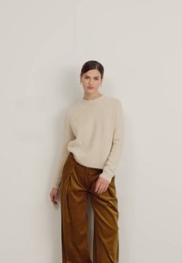 Beige Wollpullover mit geripptem Halsausschnitt, kombiniert mit weitem, strukturiertem olivgrünen Cordhosen. Das Modell steht vor einem schlichten Hintergrund.