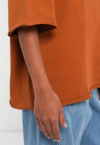 Υπερμεγέθες βαμβακερό μπλουζάκι σε χρώμα σκουριάς με κοντά μανίκια, με χαλαρή εφαρμογή και εκτεθιμένες ραφές. Εμφανίζεται δίπλα σε ανοιχτό μπλε τζιν.