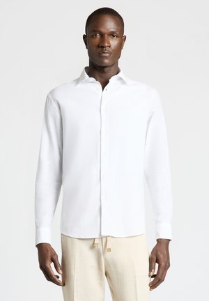 Uomo in piedi che indossa una camicia bianca a maniche lunghe abbottonata e pantaloni beige chiaro su uno sfondo chiaro e uniforme.