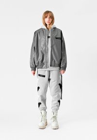 Chaqueta bomber gris con cremallera, de corte oversized, combinada con pantalones de chándal gris claro con acentos negros y detalles en patrón, llevados con botas blancas.
