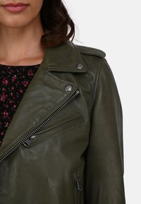 Olive green leather biker jacket with asymmetric zippers, snap buttons, and a textured finish, layered over a floral blouse.
