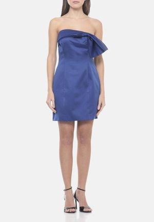 Donna che indossa un mini abito aderente in raso blu senza spalline con un grande fiocco sul petto e sandali neri con tacco alto.