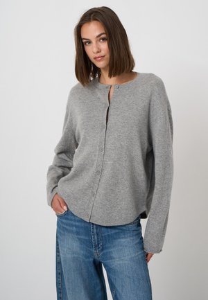 Young woman wearing a light gray button-up cardigan and blue jeans, standing against a plain light gray background.