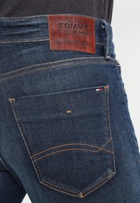 Mörkblå denimbyxor med ett brunt läderpatch som säger "TOMMY JEANS", orange sömnad och en bakficka med en liten flaggdetalj.
