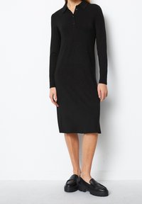 Kvinna iklädd en svart långärmad knä-lång polo klänning med svarta kraftiga loafers, stående mot en vit vägg.