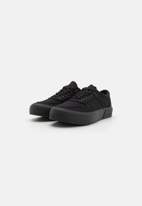 Black canvas sneakers with a textured rubber sole, flat lace closure, and reinforced stitching details. Minimalist design with a low profile.