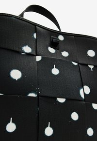 Czarna materiałowa torba o fakturowanej powierzchni, z białymi wzorami rozprysków. Posiada czarny element metalowy oraz górny uchwyt do noszenia.