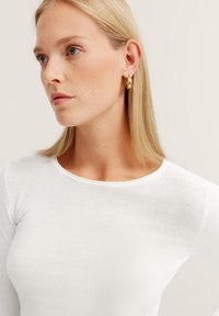 Weißes Langarmoberteil mit Rundhalsausschnitt aus glattem Stoff. Das Model hat blonde Haare und trägt goldfarbene Creolen.