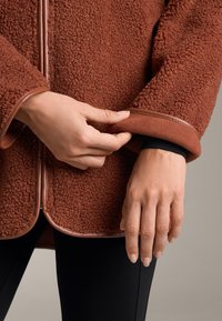 Rust-colored fleece jacket with a textured surface, trimmed in smooth brown leather. A hand adjusts the cuff, revealing a black inner layer.